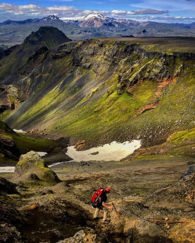A hiker descending a green volcanic ridge beneath snow-capped mountains in Iceland.