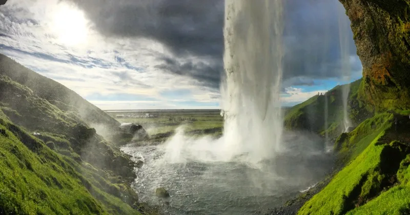 Looking out from behind a waterfall toward a sunlit valley and mist-covered rocks.