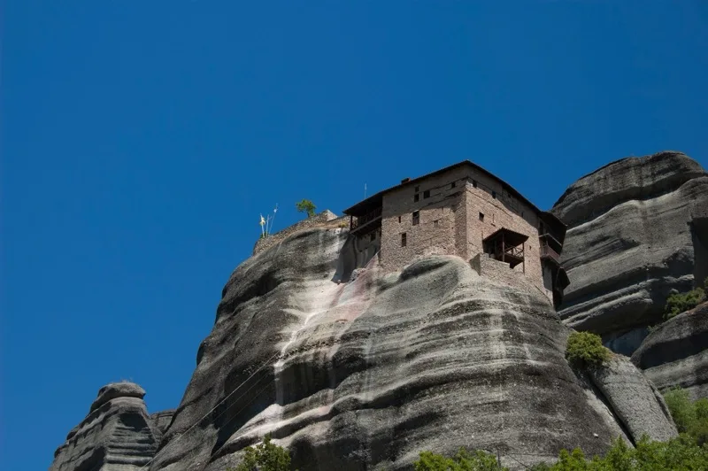 A stone monastery perched on a towering rock formation against a clear blue sky.