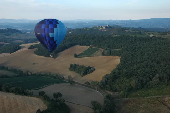 A blue hot air balloon drifting over the rolling fields and woods of the Tuscan countryside.